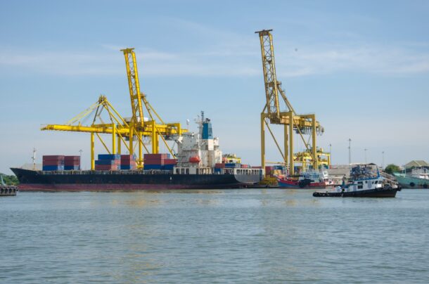 cargo ship at dock with yellow cranes, tugboat nearby on clear day