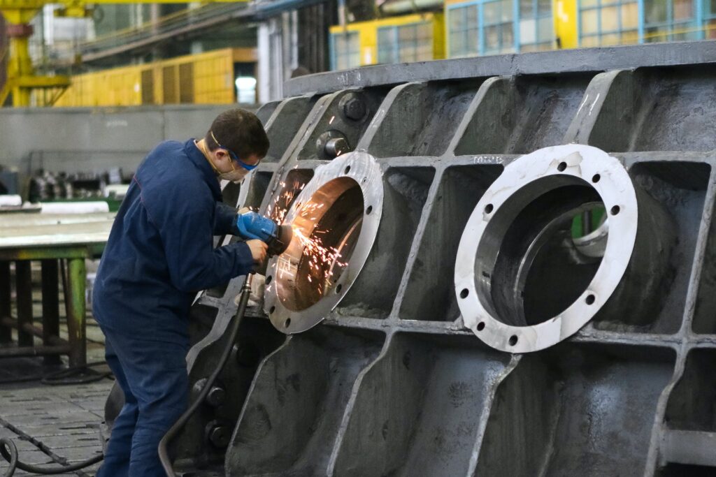 worker in blue overalls grinding a large metal part at an industrial facility