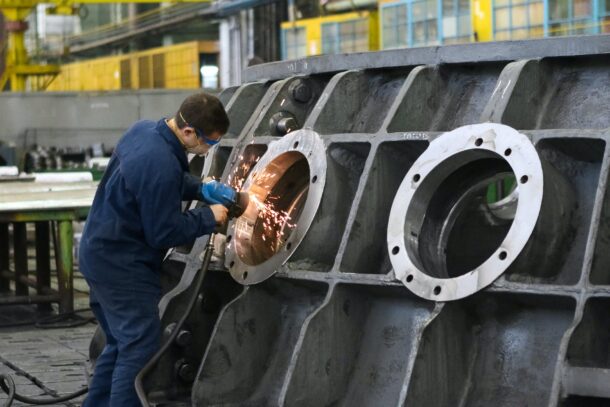 worker in blue overalls grinding a large metal part at an industrial facility